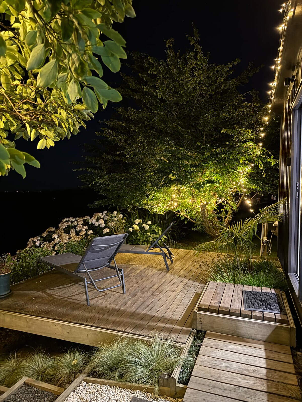 A wooden deck is illuminated by soft string lights, surrounded by lush foliage and flowering plants. Two lounge chairs are positioned for relaxation, offering serene views of the night sky and landscape beyond.