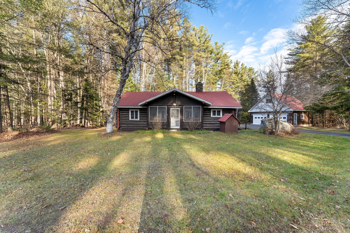 A charming log cabin is set on a spacious lawn, surrounded by tall trees. The cabin features a red metal roof and large front windows, allowing natural light inside. A detached garage is visible to the right, blending harmoniously with the natural surroundings.