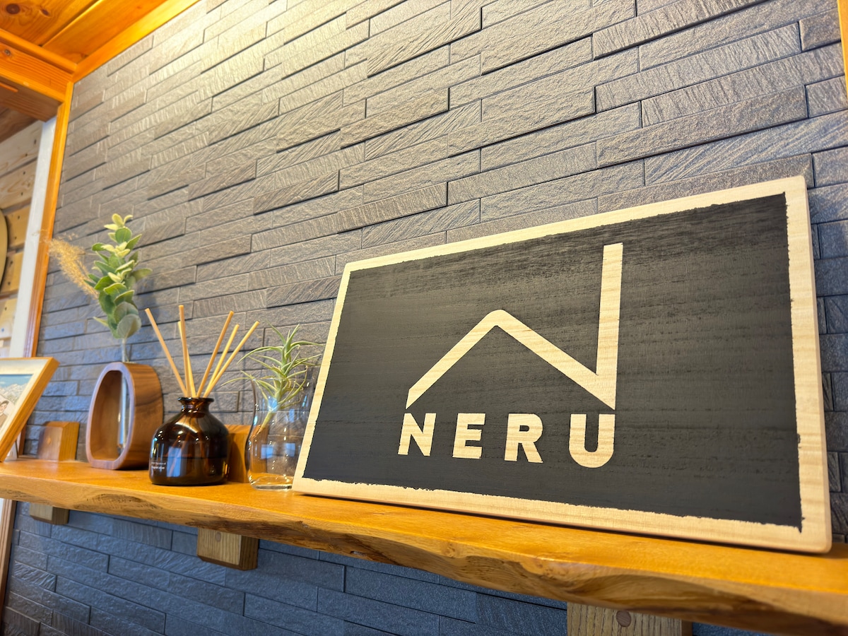 A decorative sign displaying the name 'NERU' is positioned on a wooden shelf. The background features a textured gray wall, complemented by decorative plants and a simple vase. The arrangement creates a warm, welcoming entry space.