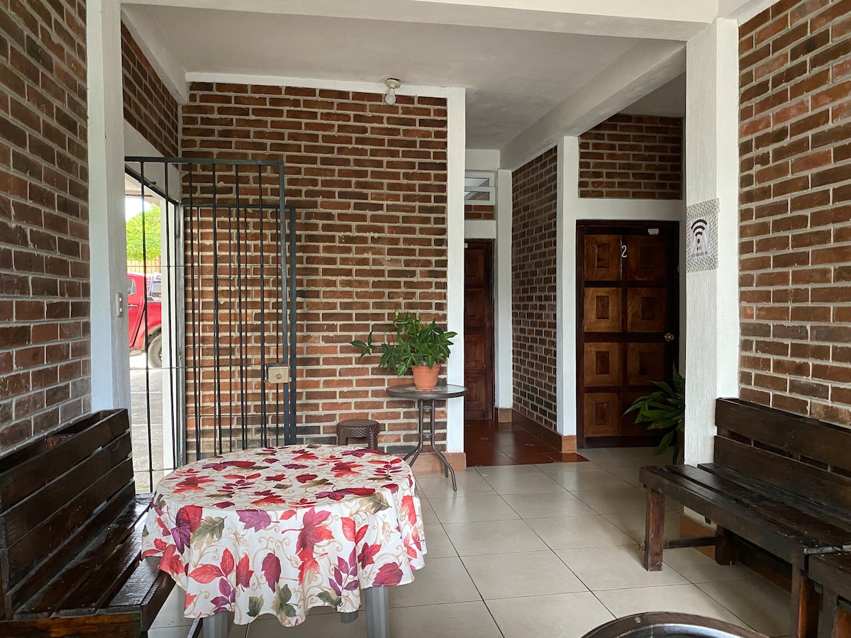 A welcoming entryway features a wooden table adorned with a floral tablecloth. The walls are lined with rustic brick, complemented by wooden benches. Potted plants add a touch of greenery, while doors to rooms create a cozy, inviting atmosphere.