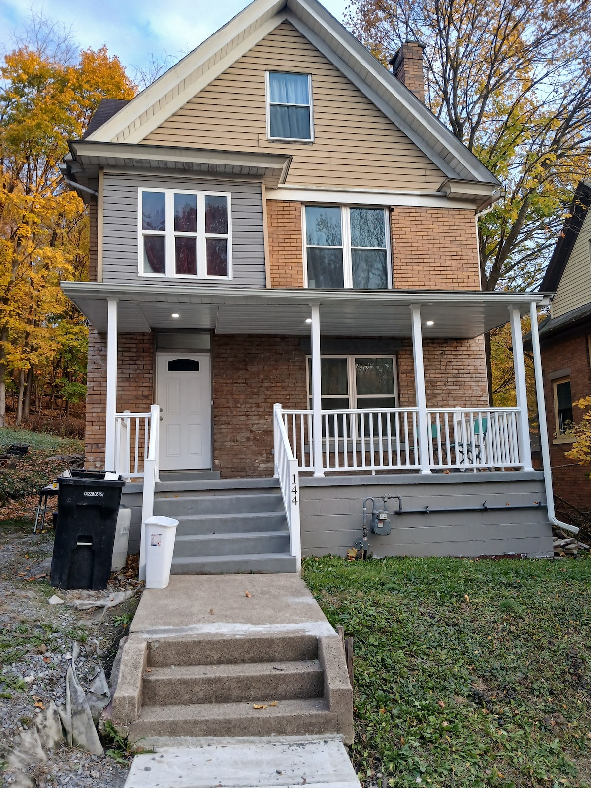 A classic three-story home features a welcoming front porch with a white railing. Large windows are set in a mix of brick and siding, allowing for ample natural light. Surrounding trees display vibrant autumn foliage, adding color to the exterior.