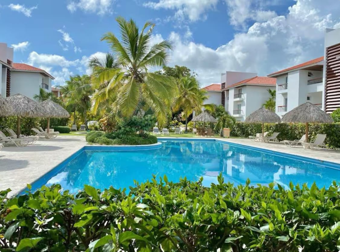 A clear blue pool is surrounded by palm trees and lounge chairs. Thatched umbrellas provide shaded areas, while well-maintained greenery frames the poolside. The bright sky is dotted with a few clouds, enhancing the inviting outdoor space.