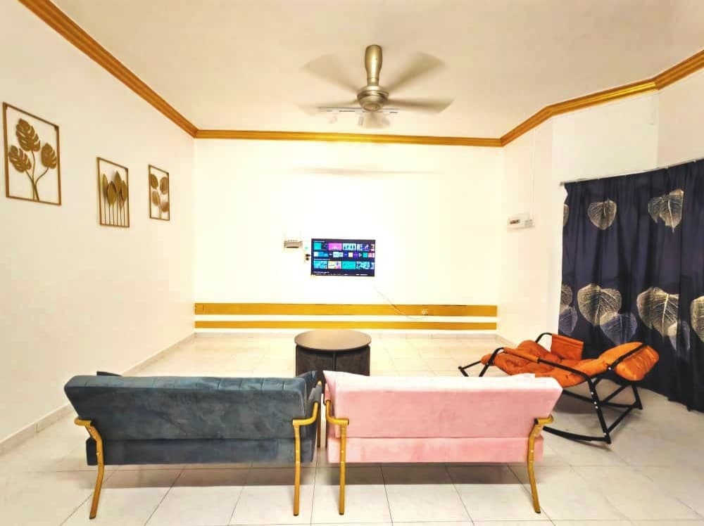 A spacious living area features two sofas in light blue and soft pink, positioned around a circular black coffee table. A wall-mounted television is visible across from the seating, and a ceiling fan provides added comfort. Dark drapes with leaf patterns frame the window.