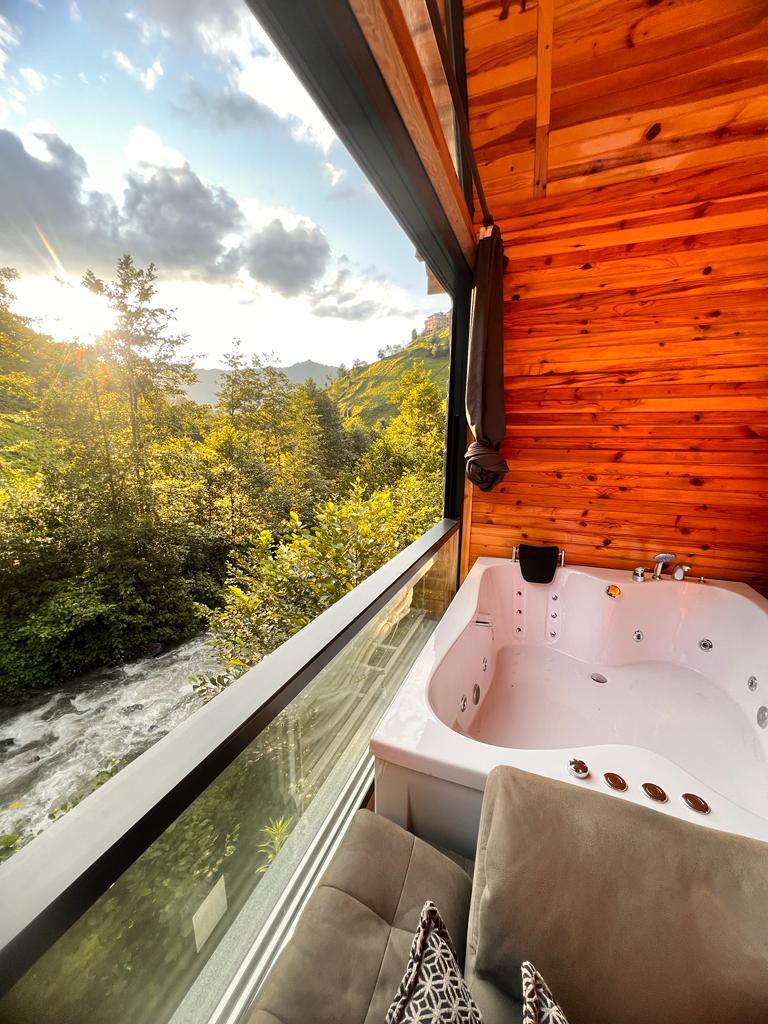 A modern hot tub is positioned next to a large window, allowing for expansive views of surrounding green trees and a flowing stream. Soft sunlight filters through the clouds, highlighting the warm wooden interior, while a cozy seating area is visible nearby.