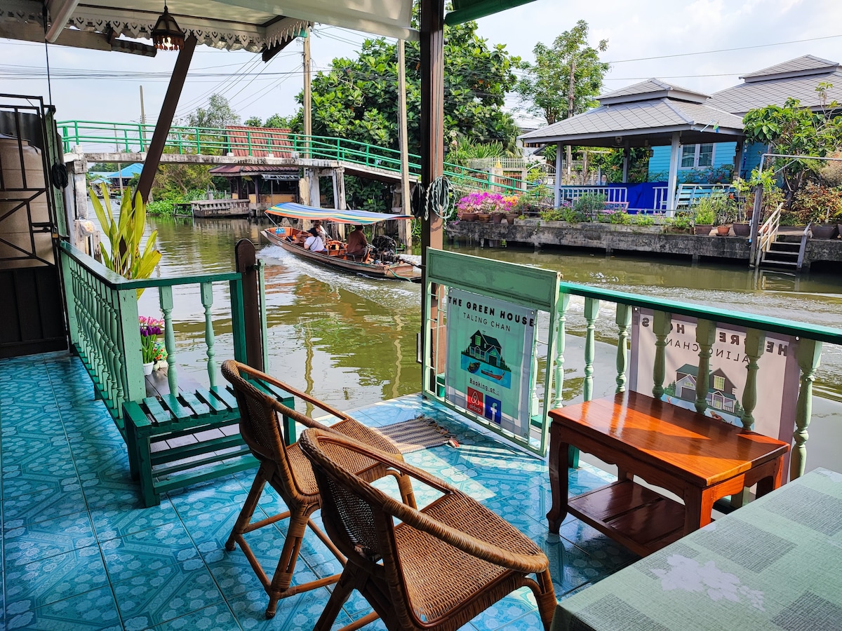 A wooden deck overlooks a serene canal, with two woven chairs positioned for relaxation. A leisurely boat passes by, complementing the tranquil water view. Lush greenery and colorful flowers adorn the edges, enhancing the calm atmosphere of the riverside setting.
