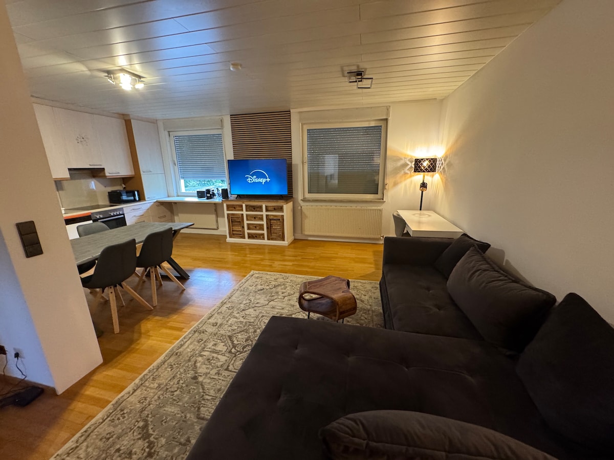 An inviting living space showcases a dark sectional couch facing a medium-sized television mounted on the wall. A dining table with light-colored chairs occupies part of the area, while a patterned rug adds texture beneath. Large windows provide natural light, complemented by a stylish lamp.