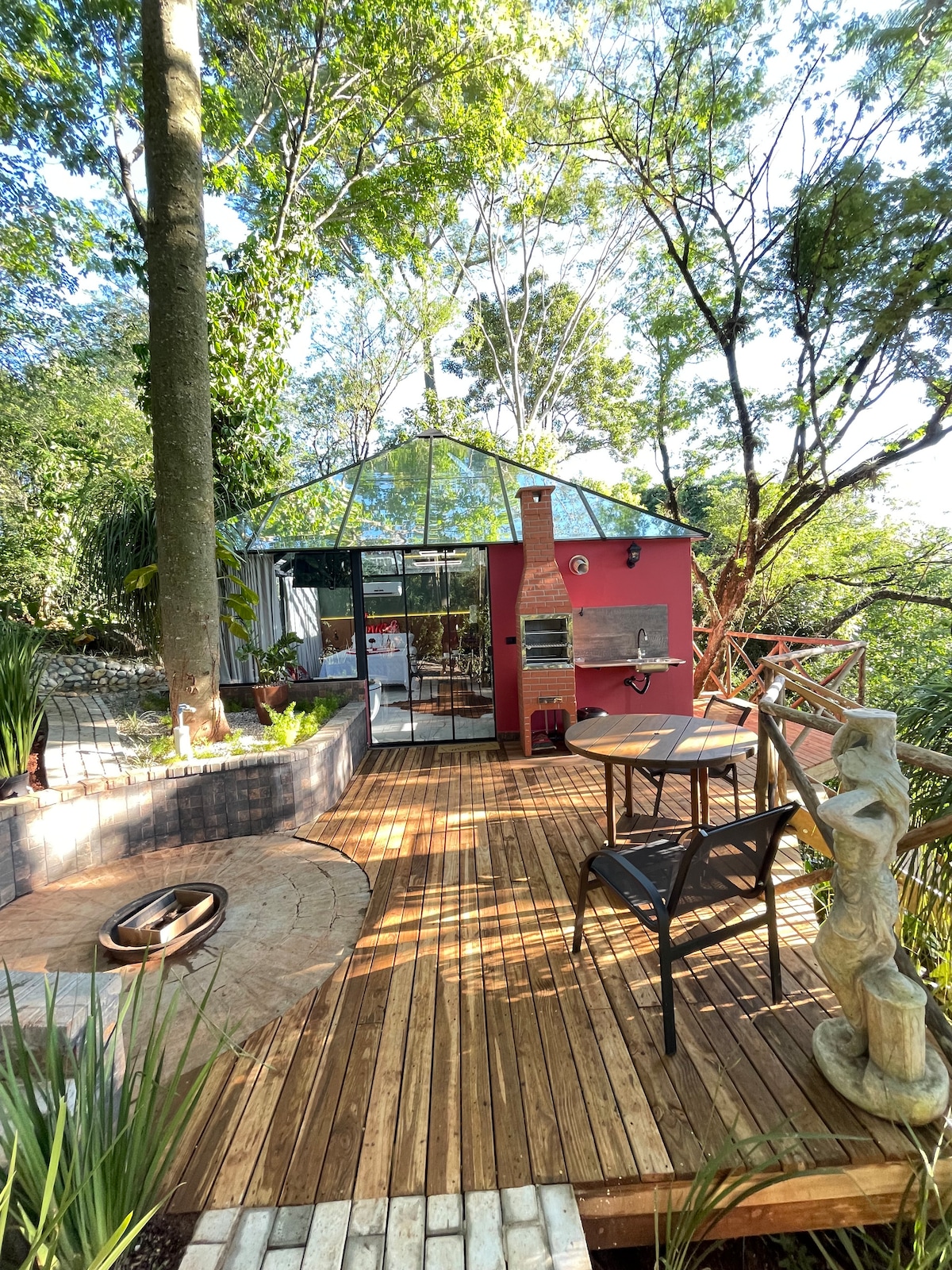 A wooden deck wraps around a vibrant red structure with large glass panels, allowing views of the surrounding greenery. A small outdoor dining set sits beside a stone sculpture and a fire pit, creating an inviting space for relaxation and outdoor gatherings.