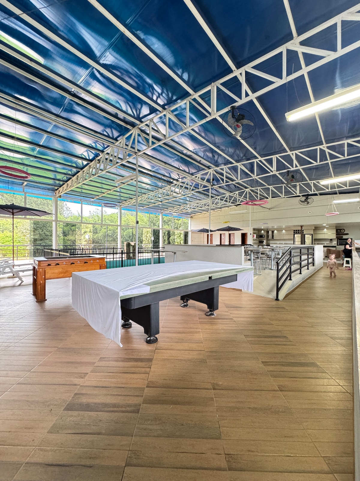 An indoor recreational area is highlighted by a large pool table covered with a white cloth. Natural light floods through expansive windows, creating a bright atmosphere. A foosball table is positioned nearby, with a staircase leading up to a lofted area visible in the background.