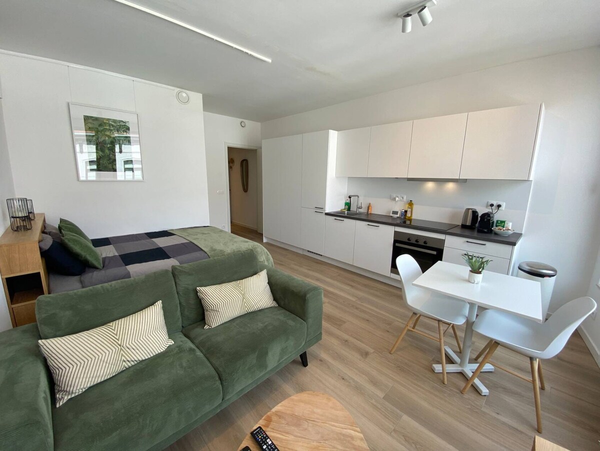 An open-plan living space features a green sofa facing a wooden coffee table. A dining area with a round table and white chairs is seen beside a modern kitchen with minimalistic cabinetry. A cozy sleeping area is included, defined by a prominent bed with a patterned throw.