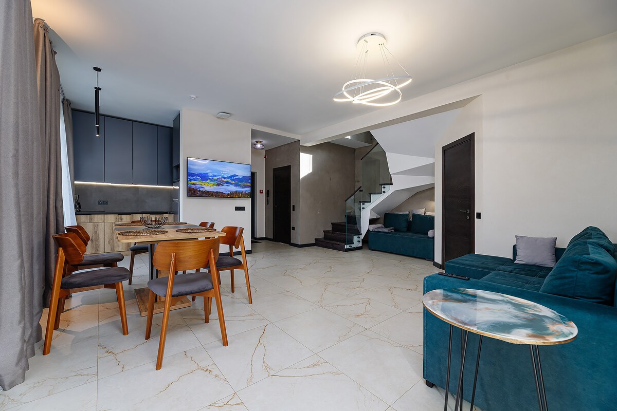 An open living area features a dining table surrounded by wooden chairs, paired with a cozy couch in a deep shade. A large screen TV is mounted on the wall, and soft lighting enhances the contemporary feel of the space.