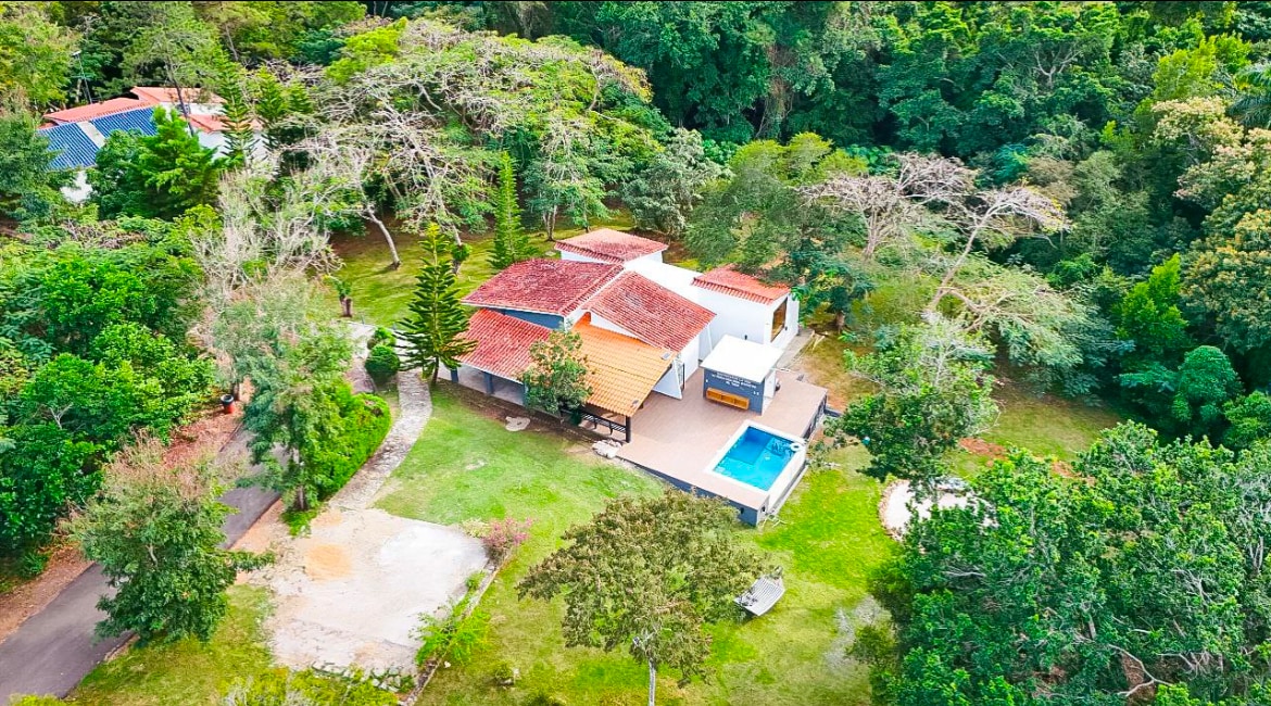An aerial view captures a spacious home nestled in lush greenery. The red-tiled roof contrasts with the vibrant surroundings. A private pool is visible in the backyard, surrounded by a well-maintained lawn, creating a serene and inviting outdoor space.