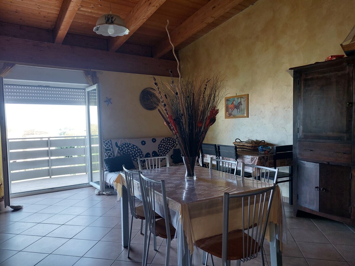 A spacious dining area features a large table surrounded by silver chairs, with a decorative arrangement in the center. Natural light enters through glass doors leading to a balcony, complemented by a wooden cabinet and artwork on the wall.