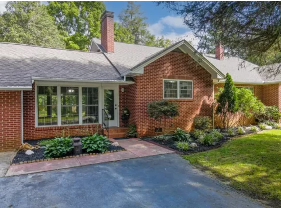 A charming brick exterior of the house is featured, with a landscaped front yard adorned with greenery. The entrance is highlighted by a porch area with large windows, allowing natural light to illuminate the interior. A paved driveway leads to the home, surrounded by soft grass.