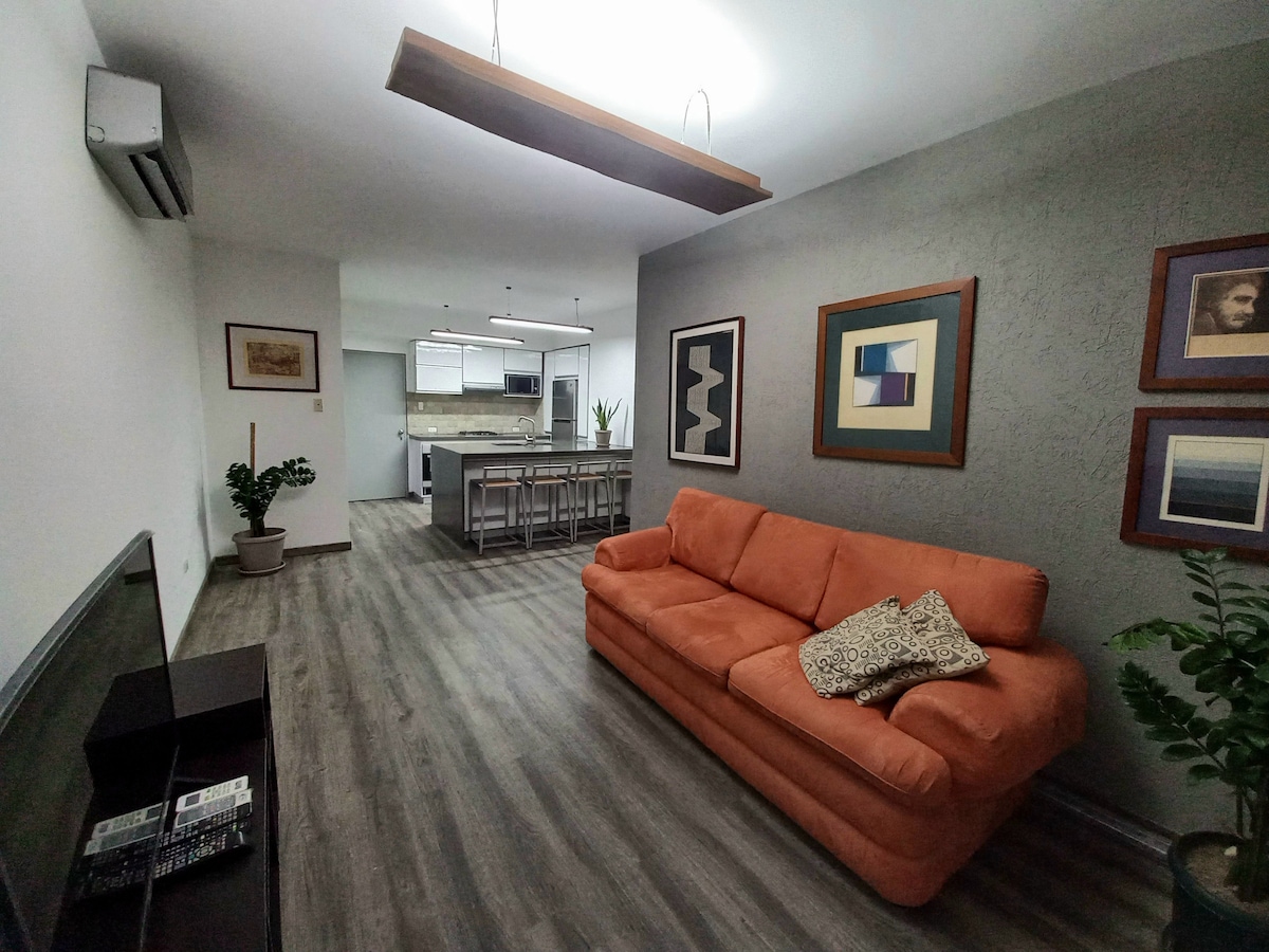 A spacious living area is furnished with an orange sofa and a decorative pillow. Artwork in frames adorns the walls, and a television sits on a dark cabinet. A modern kitchen is visible in the background, featuring a long dining table with several chairs.