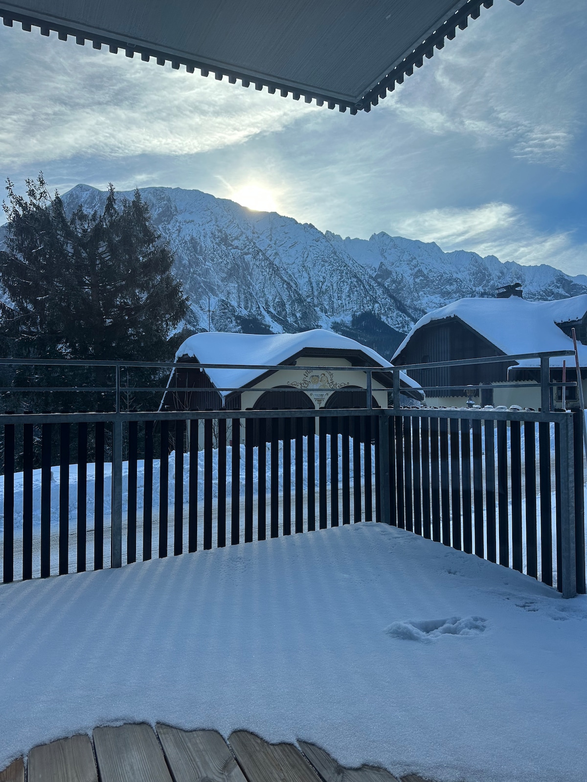 The image showcases a snowy terrace framed by a black wooden railing, offering a clear view of majestic mountains under a bright sky. The sunlight casts a gentle glow on the snow-covered landscape, emphasizing the serene winter setting.