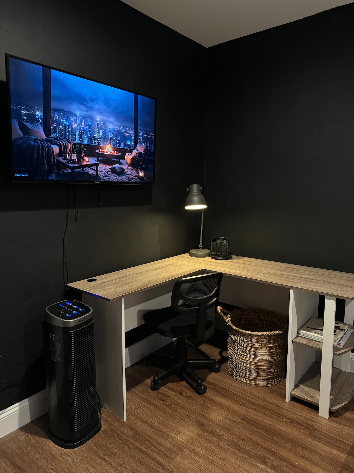 A modern workspace is featured, with a wooden corner desk and a comfortable black chair. A flat-screen TV is mounted on the dark wall, showing a cityscape. A decorative basket and a small shelf are situated nearby, along with a desk lamp and a kettle.