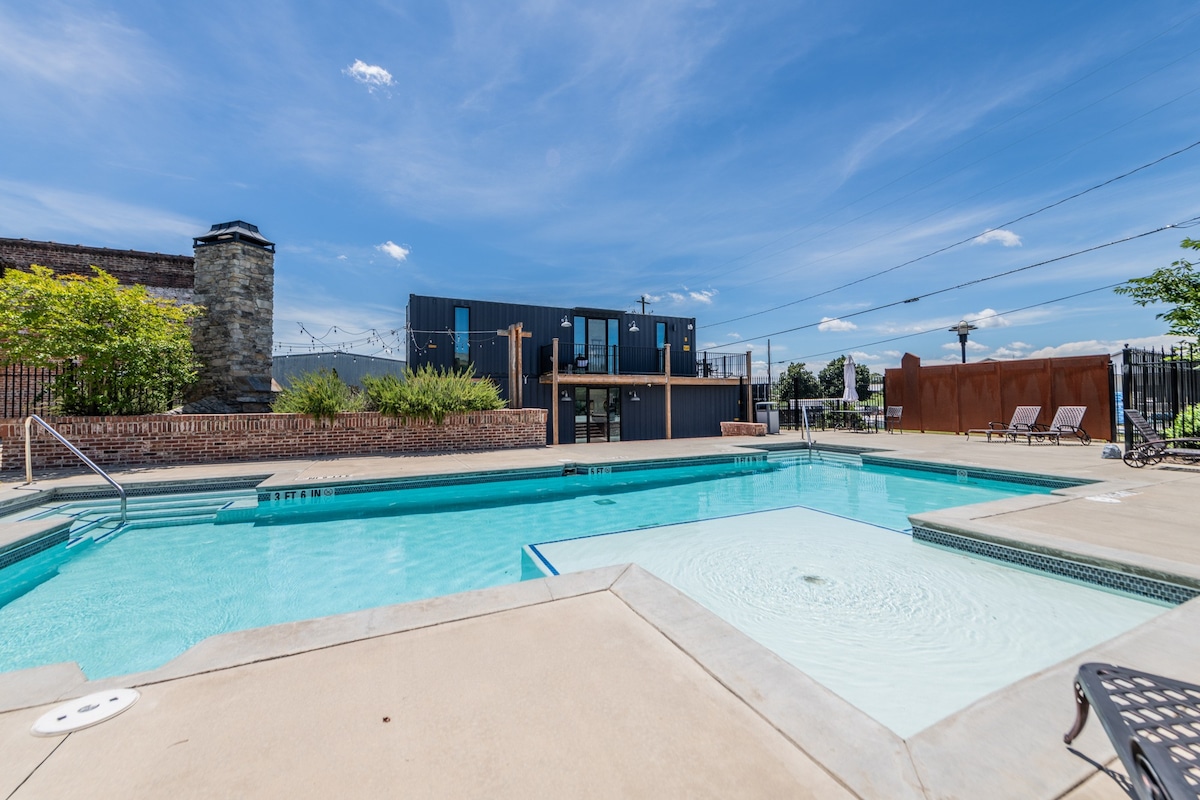 A spacious outdoor pool is surrounded by a concrete deck, featuring lounge chairs positioned nearby. Lush greenery and well-maintained landscaping are visible along the pool's edge, with a modern building in the background under a clear blue sky.