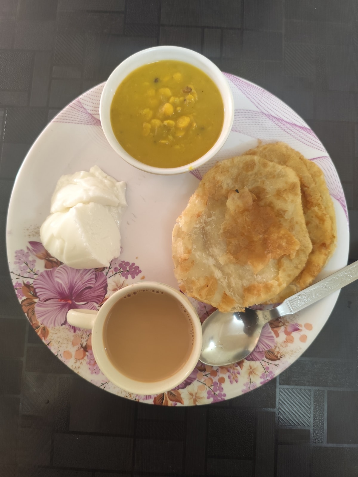 A round plate is arranged with a serving of golden-brown flatbreads alongside a bowl of lentil curry with corn. A dollop of yogurt is placed next to the breads, and a cup of tea is positioned nearby with a spoon resting on the edge.