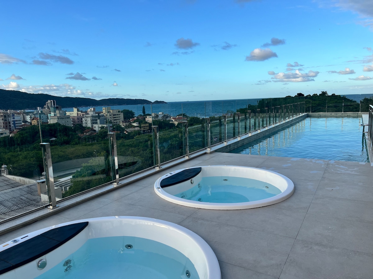 Two outdoor jacuzzis with clear waters are positioned on a spacious terrace, providing scenic views of the ocean and surrounding landscape. A large infinity pool extends nearby, framed by glass railings, with blue skies and clouds enhancing the tranquil atmosphere.