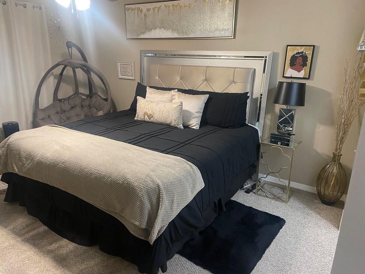 A spacious sleeping area is furnished with a large bed covered in dark bedding and an accent throw. A stylish hanging chair is positioned nearby. Decorative art and a gold-framed mirror enhance the contemporary decor, while soft lighting provides a calming ambiance.