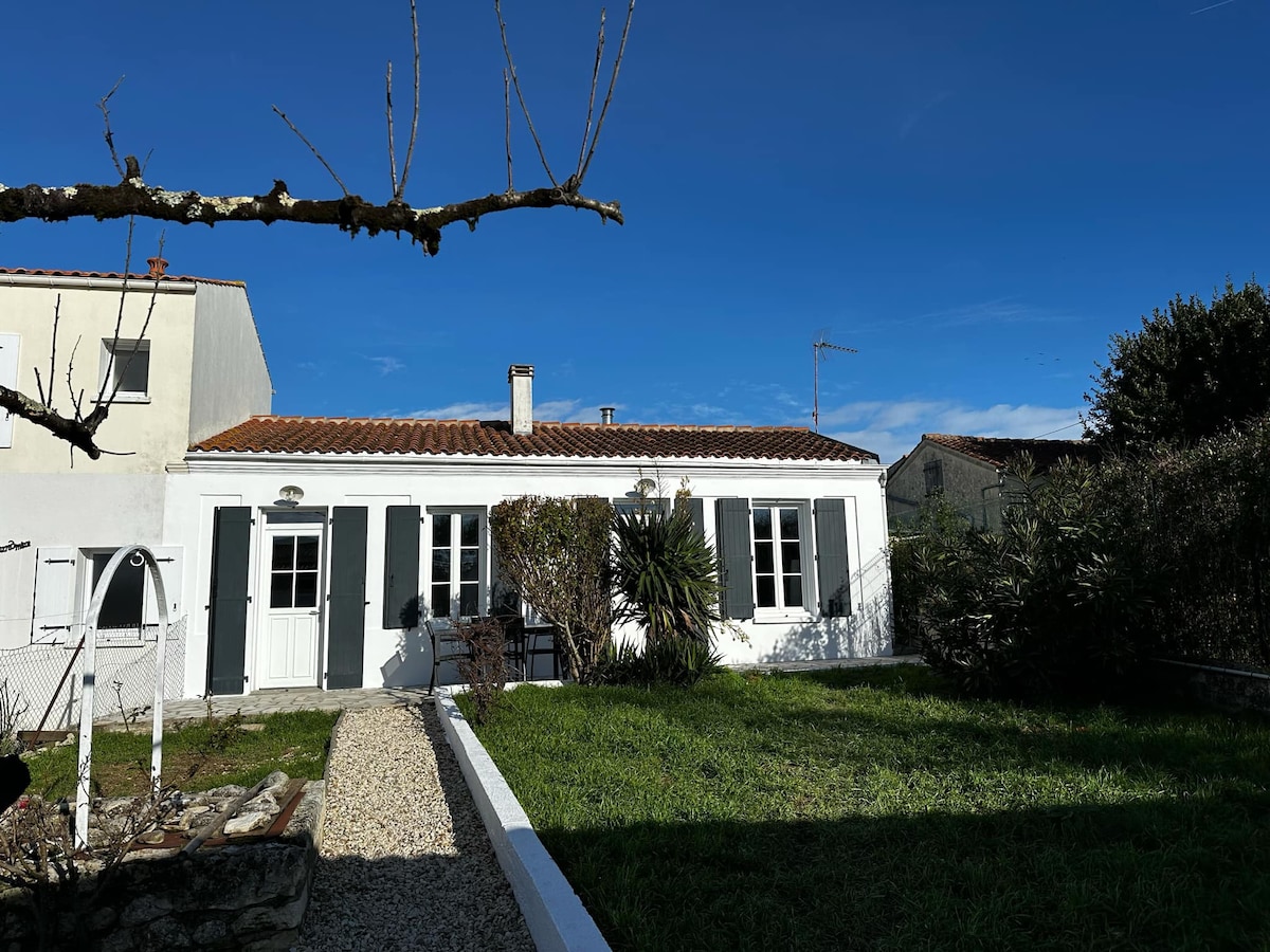 The exterior of a charming house is presented, featuring a white facade with green shutters. A spacious garden with grass is visible in the foreground, alongside a gravel pathway. The clear blue sky enhances the bright appearance of the property.