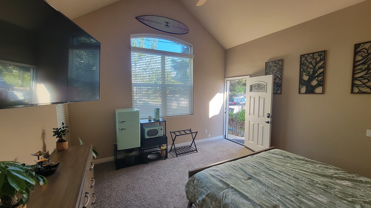 A spacious bedroom features a large window allowing natural light to enter. A flat-screen television is mounted on the wall across from the bed. A stylish dresser and a mini-fridge add functionality, while a private entrance is visible, framed by light-colored walls and accent decor.
