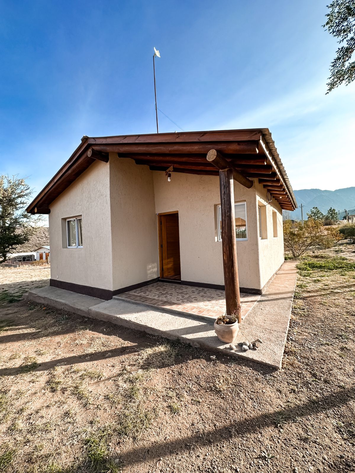 A single-story cabin is shown, featuring a peaked roof and wooden beams. Two windows allow natural light to enter, and a front porch area provides access to the entrance. The surrounding landscape includes grass and scattered trees, with mountains visible in the background.