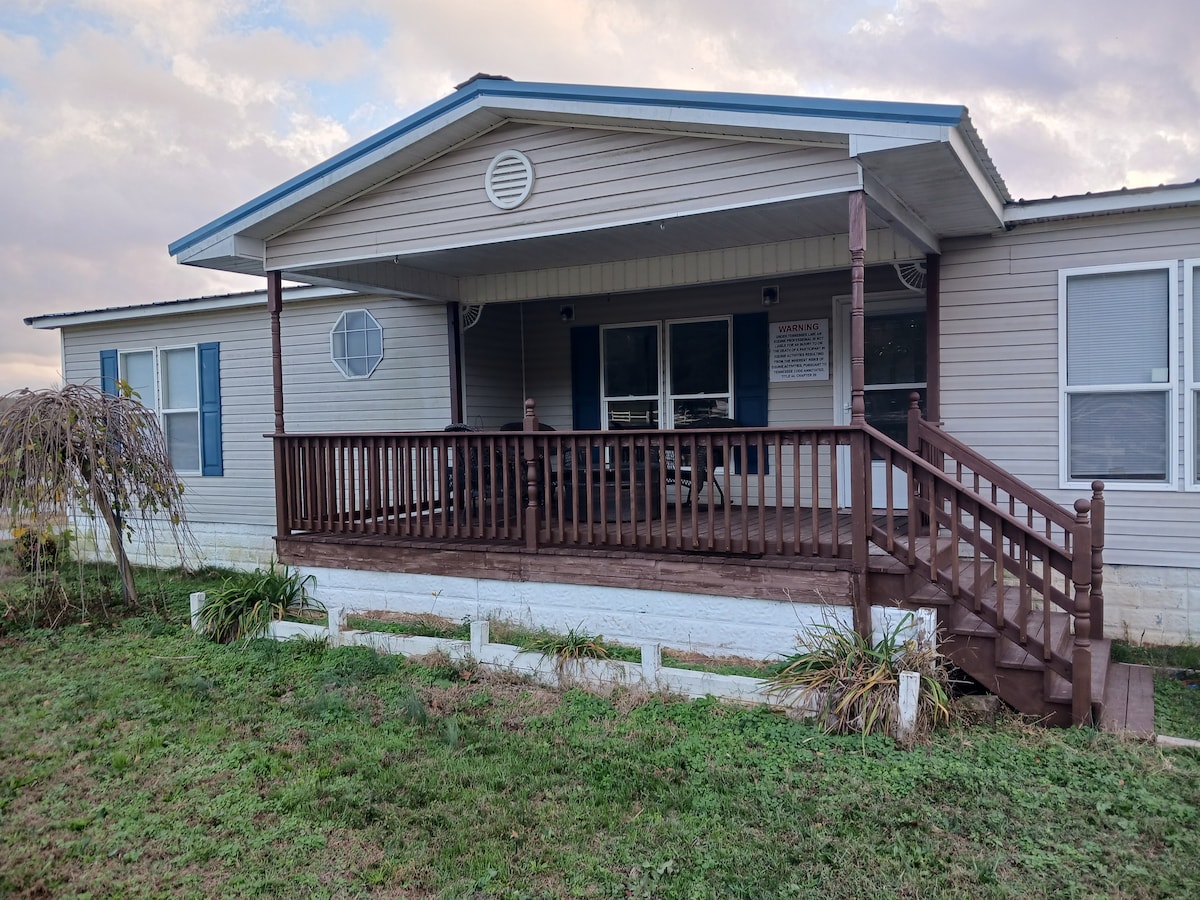 A modular home is depicted, featuring a wide front porch with wooden railings and steps. Light gray siding and a blue roof complement the warm colors of the house. Surrounding greenery adds a natural touch to the exterior setting.