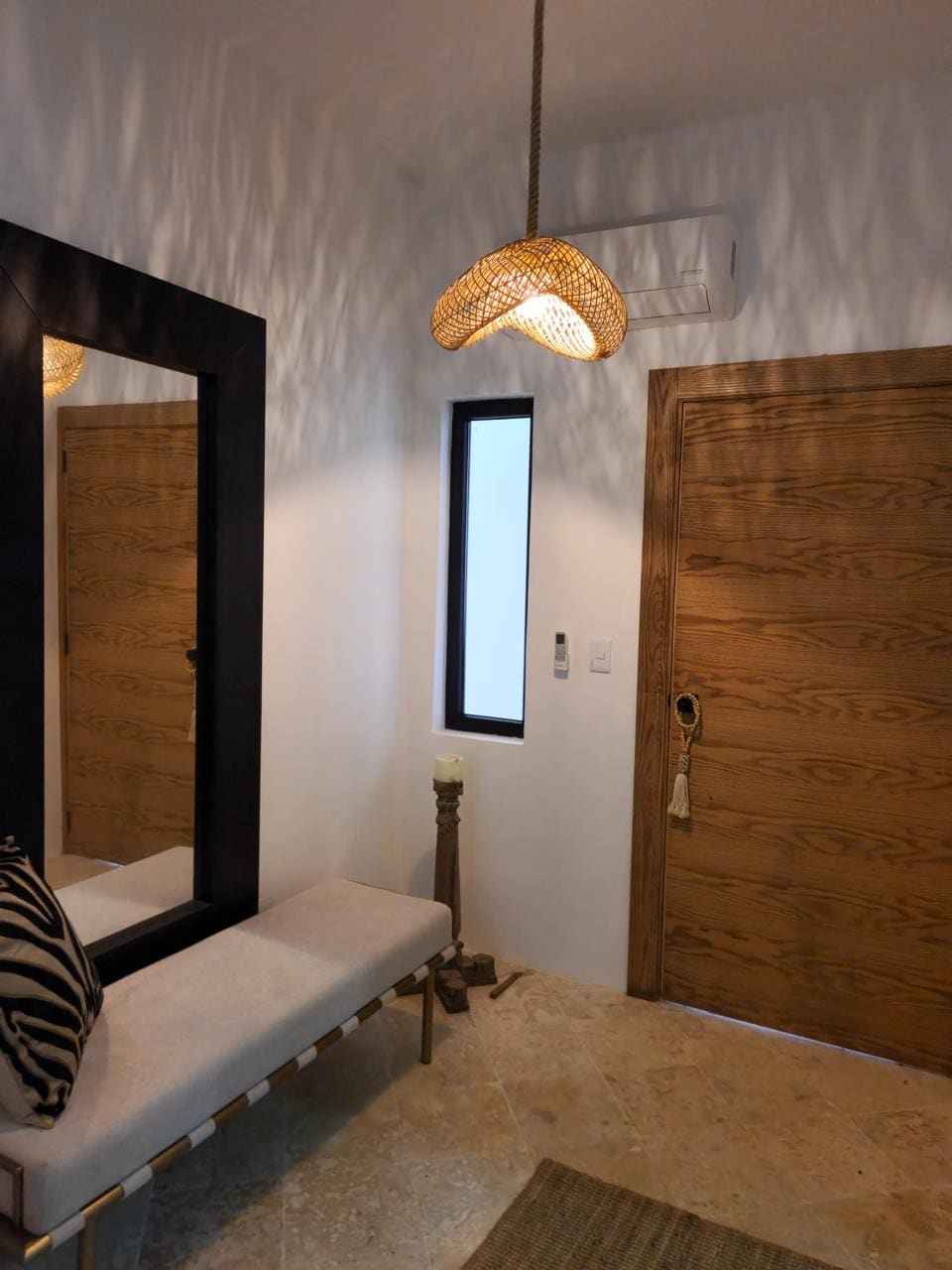 An entryway is presented with a sleek wooden door and a narrow window allowing natural light. A stylish bench with a striped cushion sits against the wall, accompanied by a full-length mirror. A woven pendant light hangs softly above, casting delicate shadows.