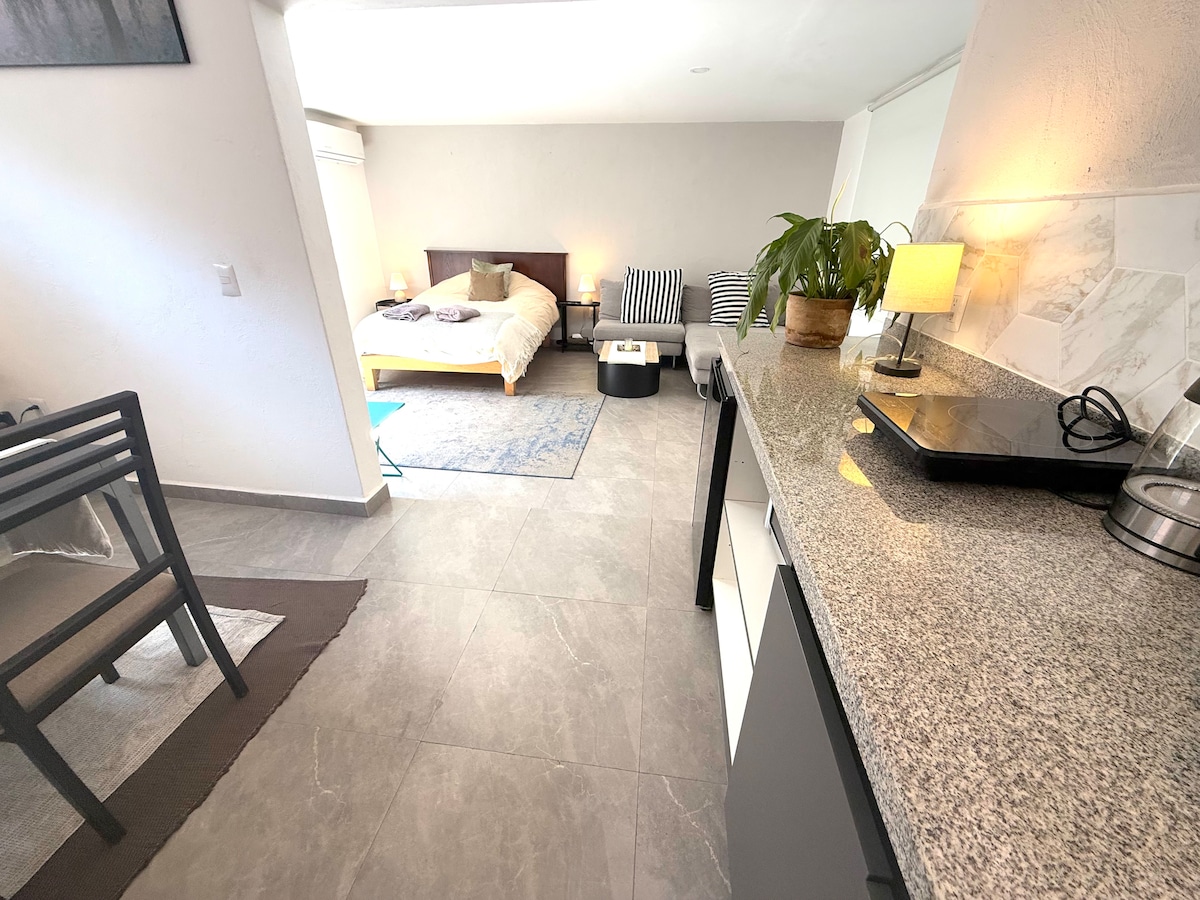 An open space is presented featuring a well-equipped kitchen with granite countertops. A cozy sleeping area includes a spacious bed, complemented by seating arrangements and decor. Natural light enters through a window, illuminating the area and promoting a bright and airy ambiance.
