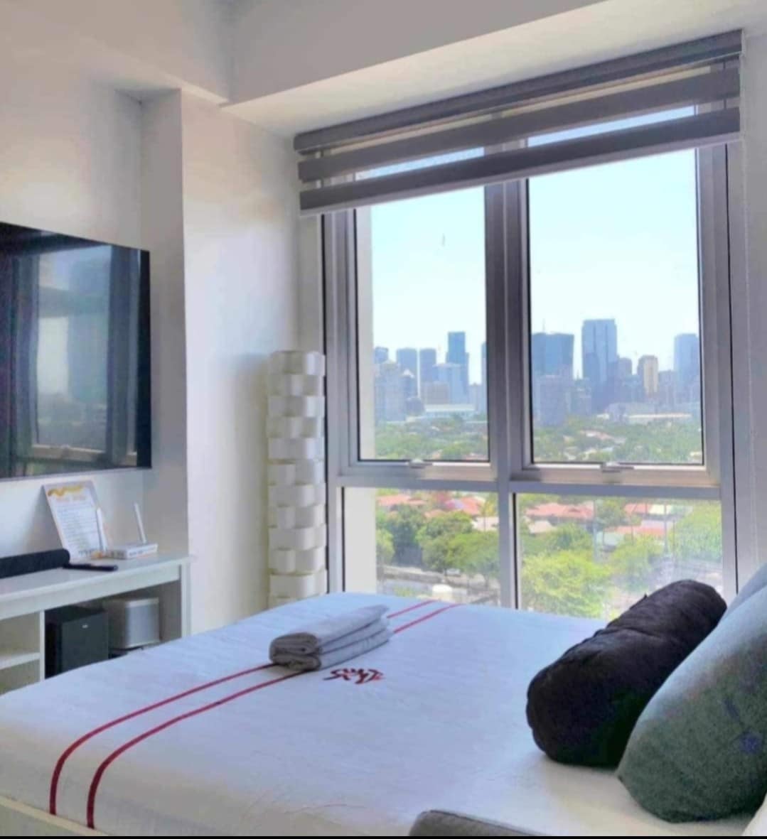 A well-lit bedroom features a spacious bed adorned with soft linens and accent pillows. Large windows offer views of the city skyline and allow natural light to fill the room. A sleek television is mounted on the wall opposite, enhancing the modern design.