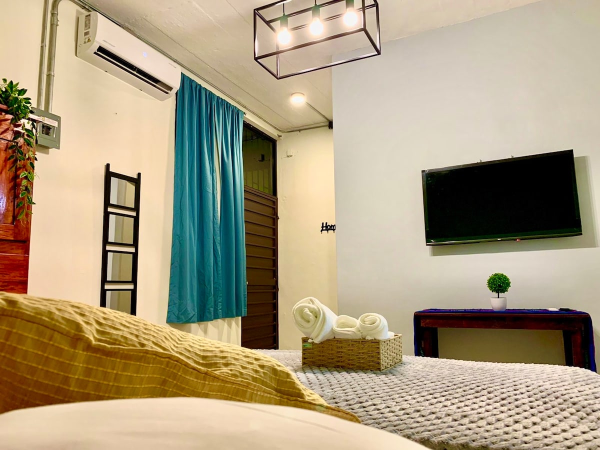A cozy sleeping area is highlighted by a soft, textured blanket draped over the spacious bed. A wall-mounted television is mounted opposite a small wooden table. Fresh towels are neatly arranged in a woven basket, and curtains gently frame a nearby window.