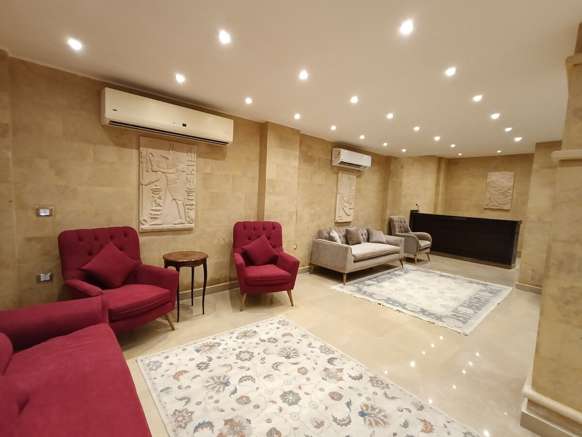 A spacious living area is furnished with two red armchairs and a gray sofa. A small wooden table is placed between the chairs. The room features textured stone walls and is well-lit by recessed ceiling lights. A decorative rug lies on the floor.