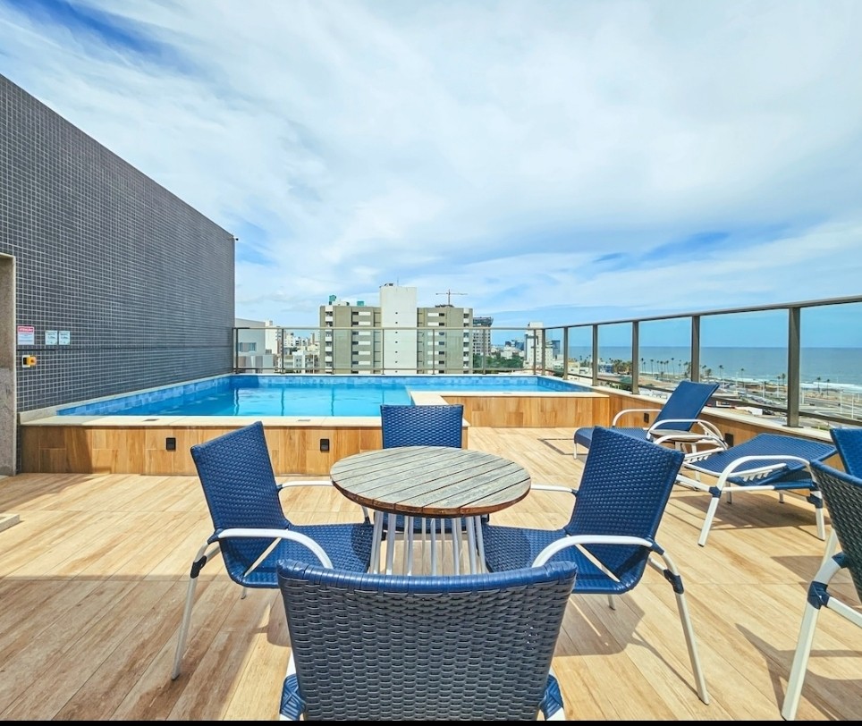 An elevated rooftop space features a swimming pool with clear blue water, surrounded by wooden decking. Several blue lounge chairs and a round table are positioned for relaxation, with a panoramic view of the skyline and ocean in the background.