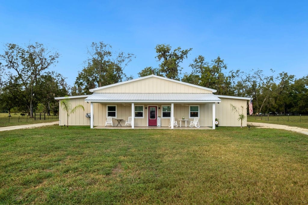 A spacious ranch-style building is framed by a lush green lawn. Two white rocking chairs are placed on the covered porch, inviting relaxation. Large windows offer views of the surrounding trees, while a prominent purple door adds a touch of color to the exterior.