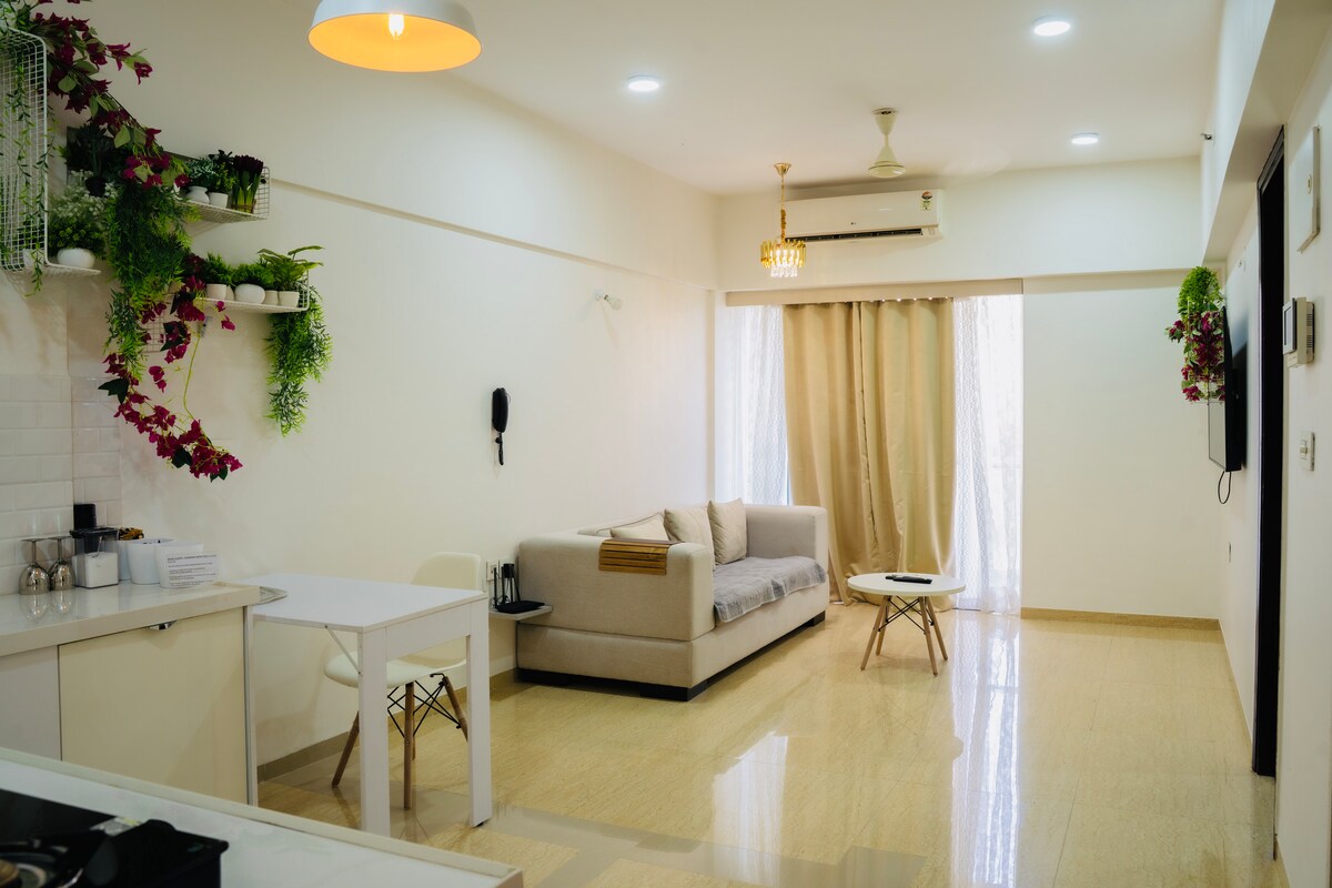 A bright living area features a luxurious sofa paired with a light-colored coffee table. Natural light filters through sheer curtains, highlighting the modern aesthetic. Greenery adorns the shelves, adding a fresh touch to the neutral-toned space.