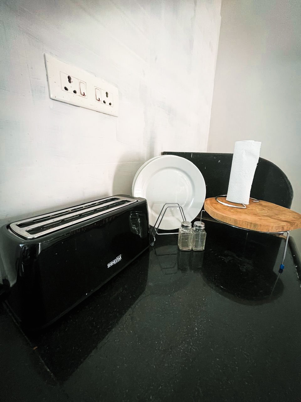 A neatly arranged kitchen counter features a black toaster, a round wooden cutting board, and a pair of glass salt and pepper shakers. A stack of plates and a paper towel holder are also included, all set against a minimalistic backdrop.