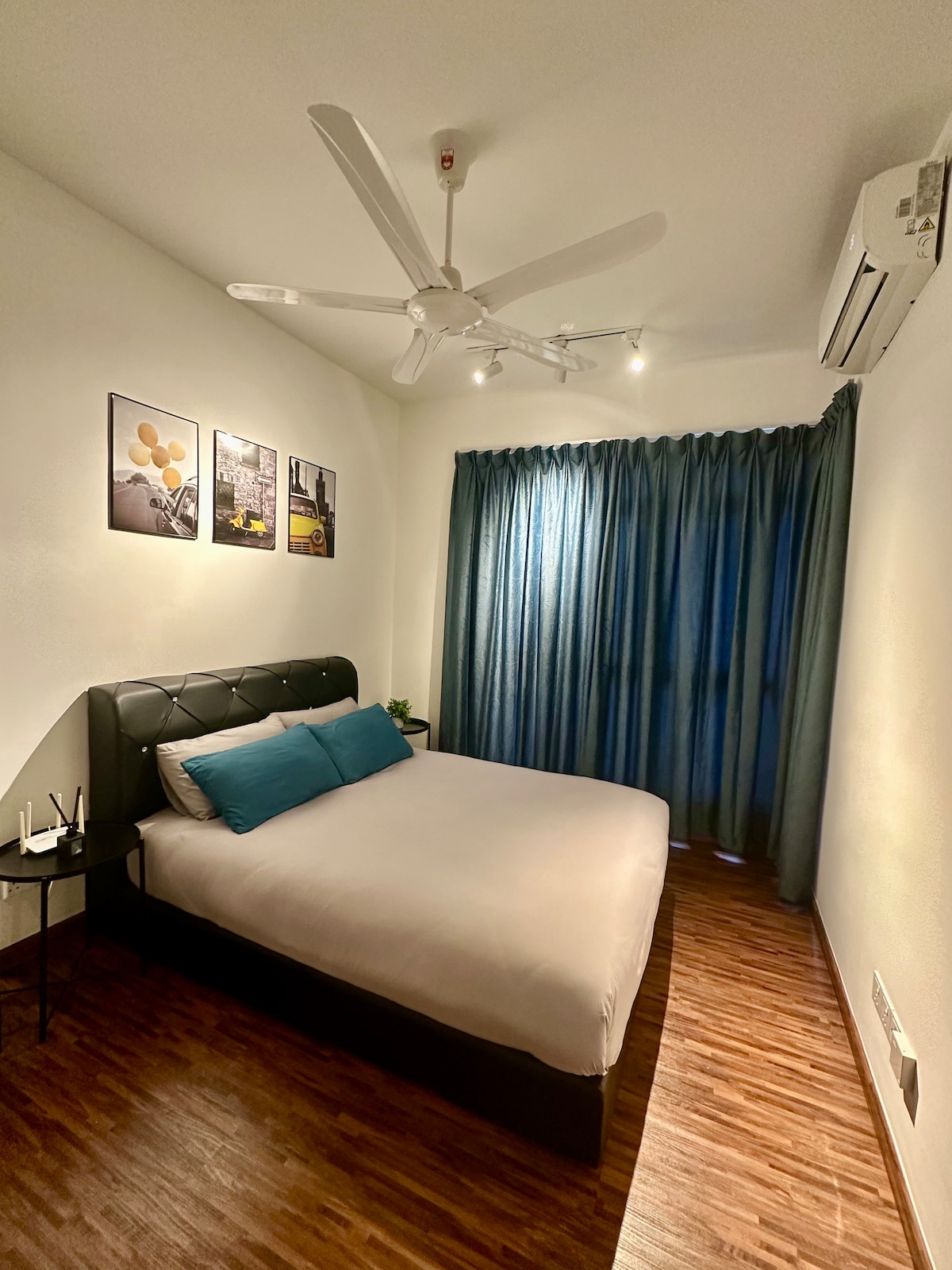 A bright bedroom features a spacious bed with decorative pillows and complemented by a stylish headboard. Artwork hangs on the walls, while a ceiling fan provides added comfort. Soft curtains frame a window, allowing natural light to fill the room, with wooden flooring adding warmth.