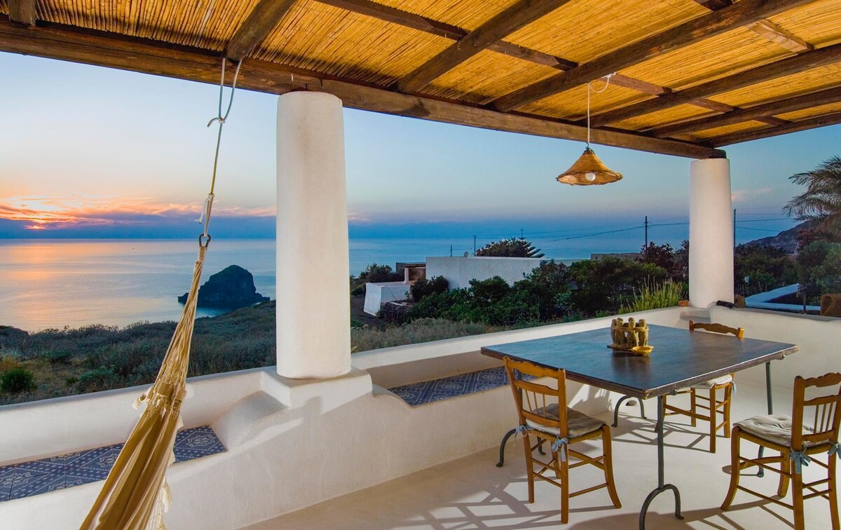 An outdoor terrace features a long dining table surrounded by wooden chairs, offering an unobstructed view of the sea. A hammock is suspended nearby, and a pendant light hangs from the ceiling, creating a serene space to enjoy sunset hues over the water.