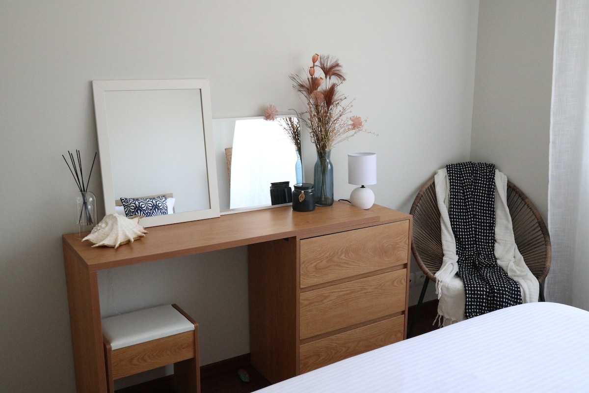 A well-lit bedroom corner features a wooden dresser with multiple drawers, accompanied by a simple white chair. A framed mirror rests on the dresser alongside decorative elements including dried plants and a small lamp. A cozy chair with a patterned throw complements the space.
