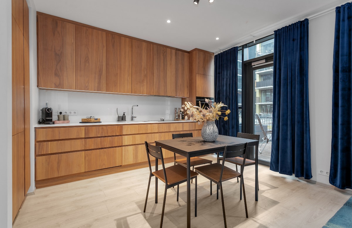 A modern kitchen showcases smooth, wooden cabinetry paired with minimalist black handles. A dining table is surrounded by four chairs, offering a comfortable setting. Natural light filters in through large windows, complemented by rich blue curtains draping beside them.