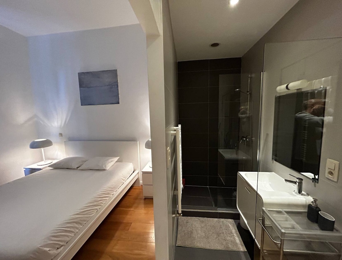 A bright sleeping area features a spacious bed framed by two minimalist nightstands with lamps. Adjacent to the sleeping area, a modern bathroom is visible, showcasing tiled surfaces, a glass shower, and a sleek sink with a large mirror.