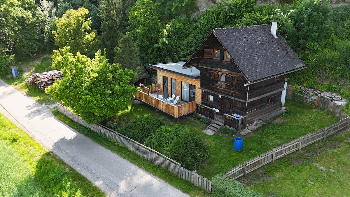 An aerial view of a rustic wooden house surrounded by greenery, showcasing two distinct sections: the original dark wood structure and a modern light wood extension. A spacious terrace with seating is visible in the front, alongside well-kept gardens and a nearby road.