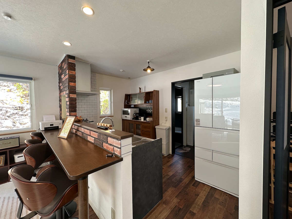The interior space features a modern open-concept design, showcasing a sleek kitchen with white cabinetry and a spacious bar area with curved stools. Natural light filters through windows, illuminating the warm wood flooring and brick accent wall.