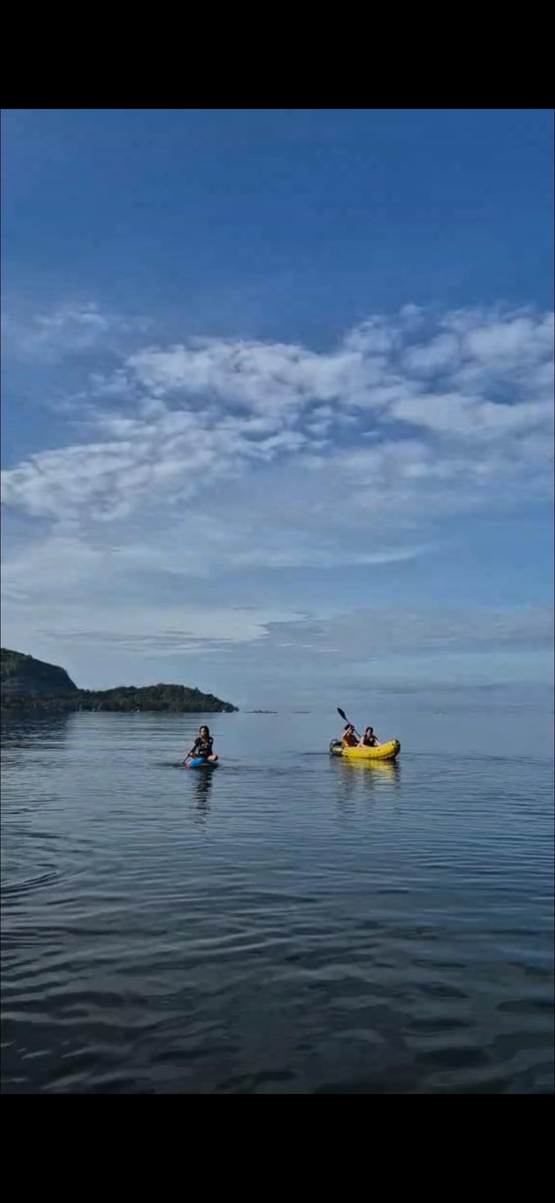 Two individuals are seen kayaking on calm waters, surrounded by gentle waves and a clear sky. One person is paddling a blue kayak, while another enjoys a bright yellow kayak. The landscape features small hills in the background, creating a serene aquatic environment.