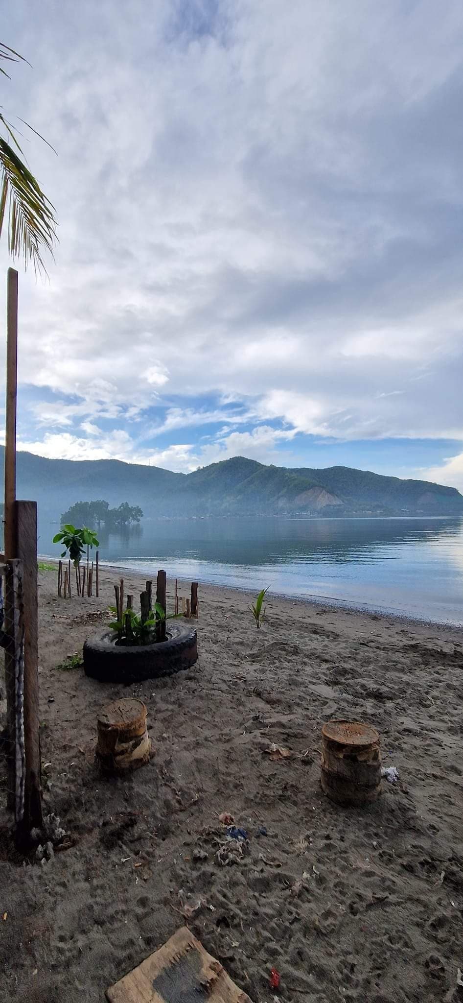 A sandy beach stretches towards calm waters, framed by lush mountains in the background. Tire planters and wooden stumps add rustic touches to the scene. A gentle sky transitions from gray to softer blue, enhancing the tranquil ambiance of the beachfront setting.