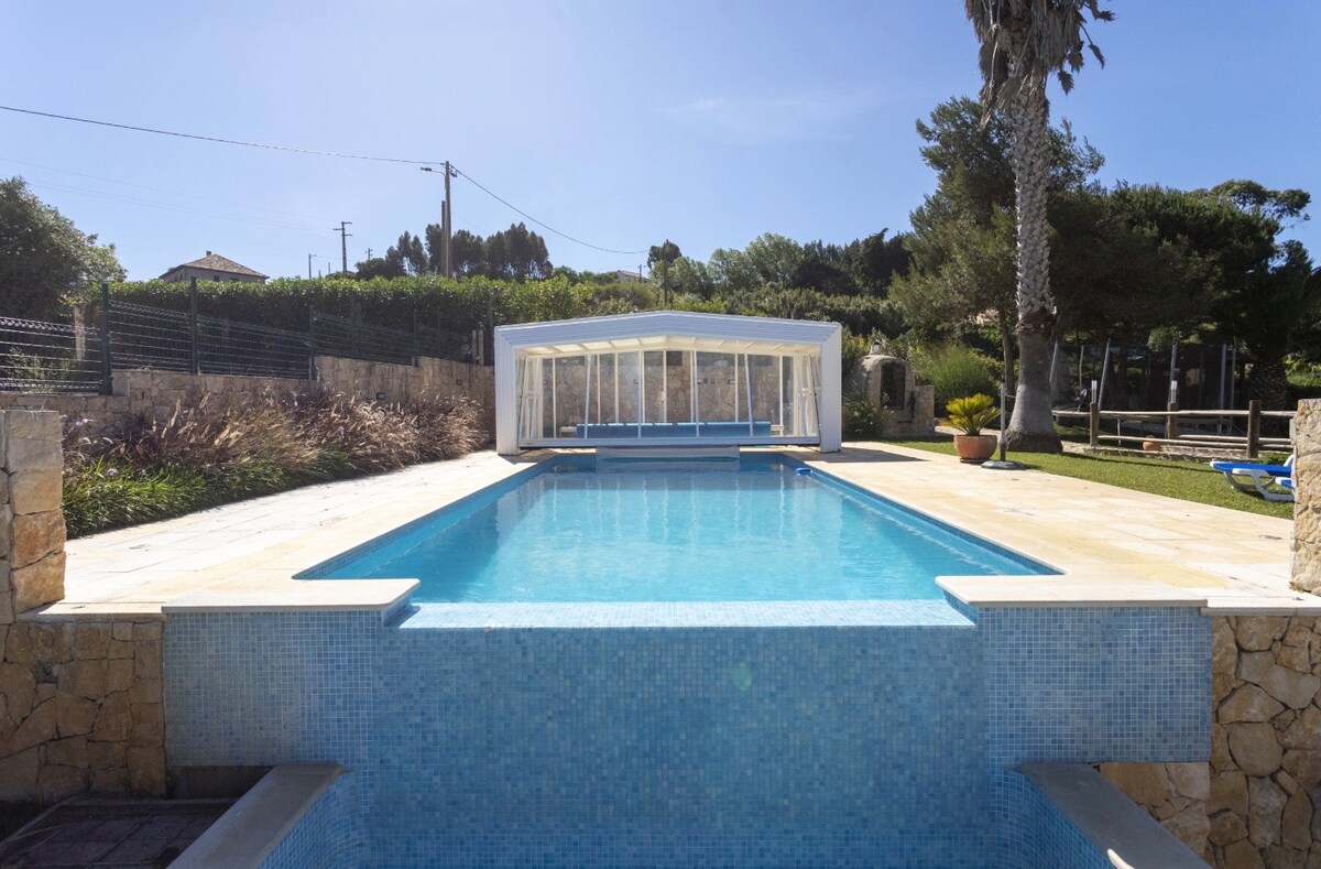 A clear and inviting swimming pool is framed by clean stone edging. The pool area is surrounded by lush greenery and features a pool house in the background. Sunlight reflects off the water's surface, enhancing a sense of tranquility in the outdoor space.