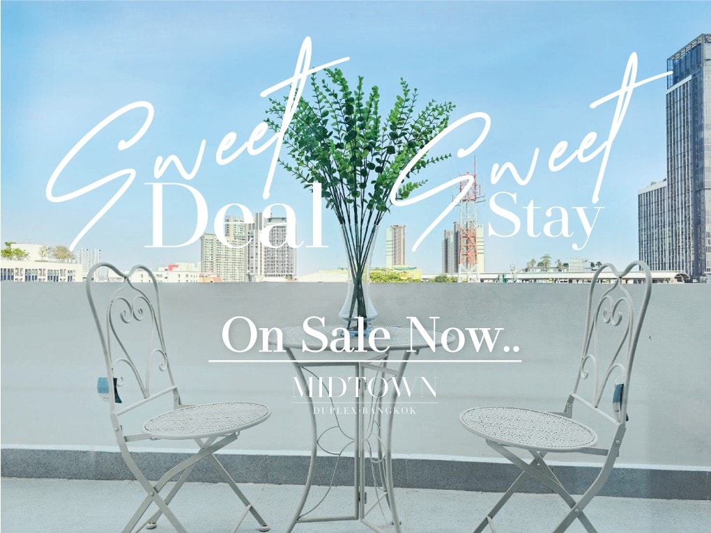 A balcony is shown, featuring two elegant white chairs positioned around a small table. Lush greenery is displayed in a vase at the center. The city skyline is visible in the background, offering a glimpse of surrounding buildings.