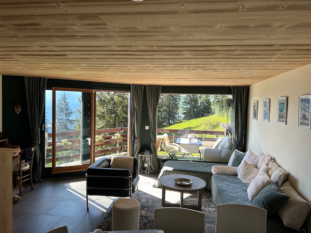 The living area features large windows framing a picturesque view of the outdoors. A mix of seating options is present, including a spacious sofa and a leather armchair. Sunlight floods the room, highlighting the wooden ceiling and the contemporary decor.