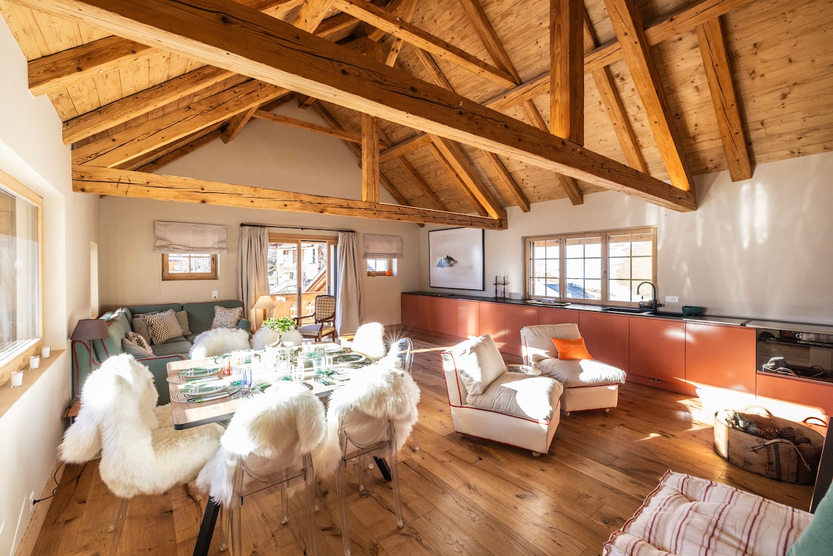 The spacious dining area features a wooden ceiling with exposed beams, complemented by natural light from large windows. A table is set for dining, surrounded by comfortable chairs covered in soft materials. A cozy seating area with plush furnishings invites relaxation.
